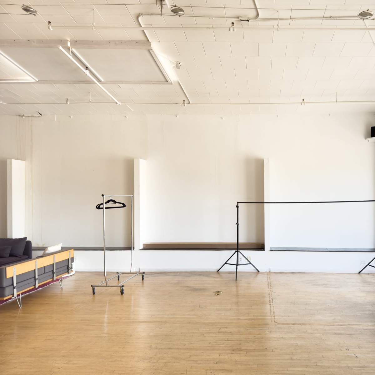 An empty interior space features a wooden floor, a large window, a clothing rack, and a simple stage area.