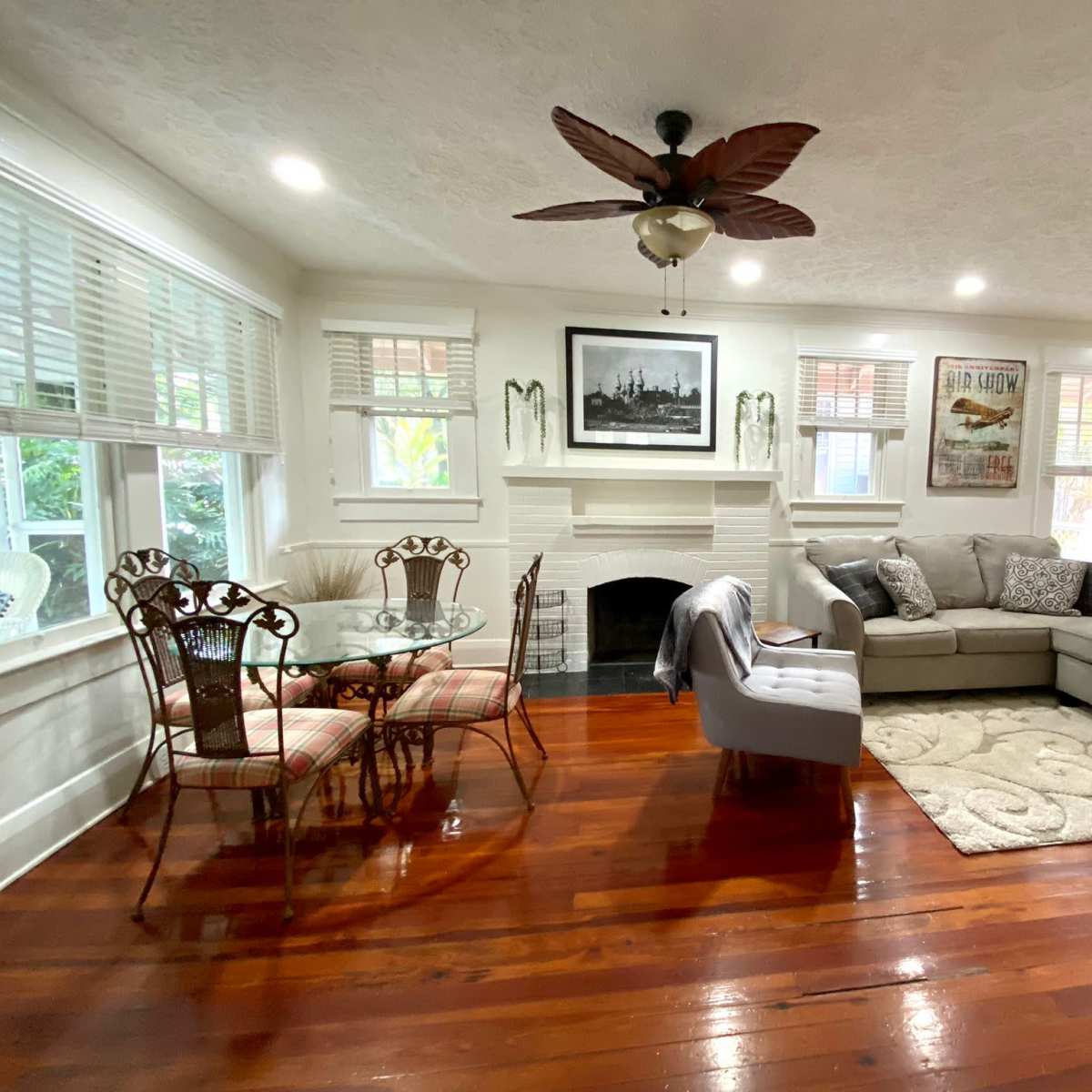 A cozy living area with a sectional sofa, a coffee table, a dining set, and a fireplace, all featuring polished wooden floors and bright windows.