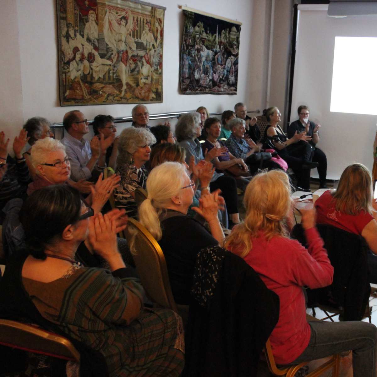 A group of seated audience members applauds as a performer stands on stage in front of a projection screen.