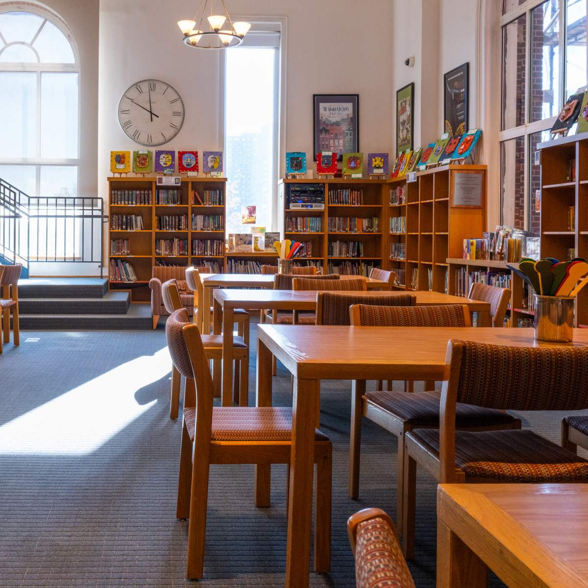 A well-lit library features wooden tables and chairs, surrounded by bookshelves filled with colorful books, under large windows and a clock on the wall.