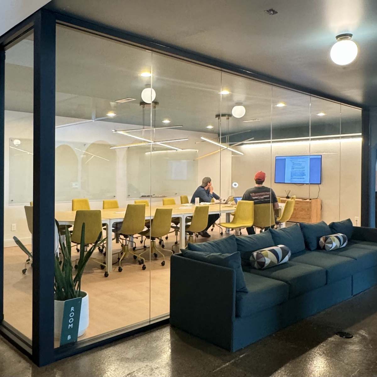 A modern conference room with glass walls features a long table surrounded by yellow chairs, while two people work at a computer in the foreground.