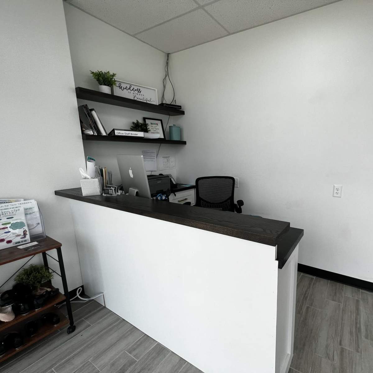 A minimalist reception desk with a computer and books on a shelf against a white wall.