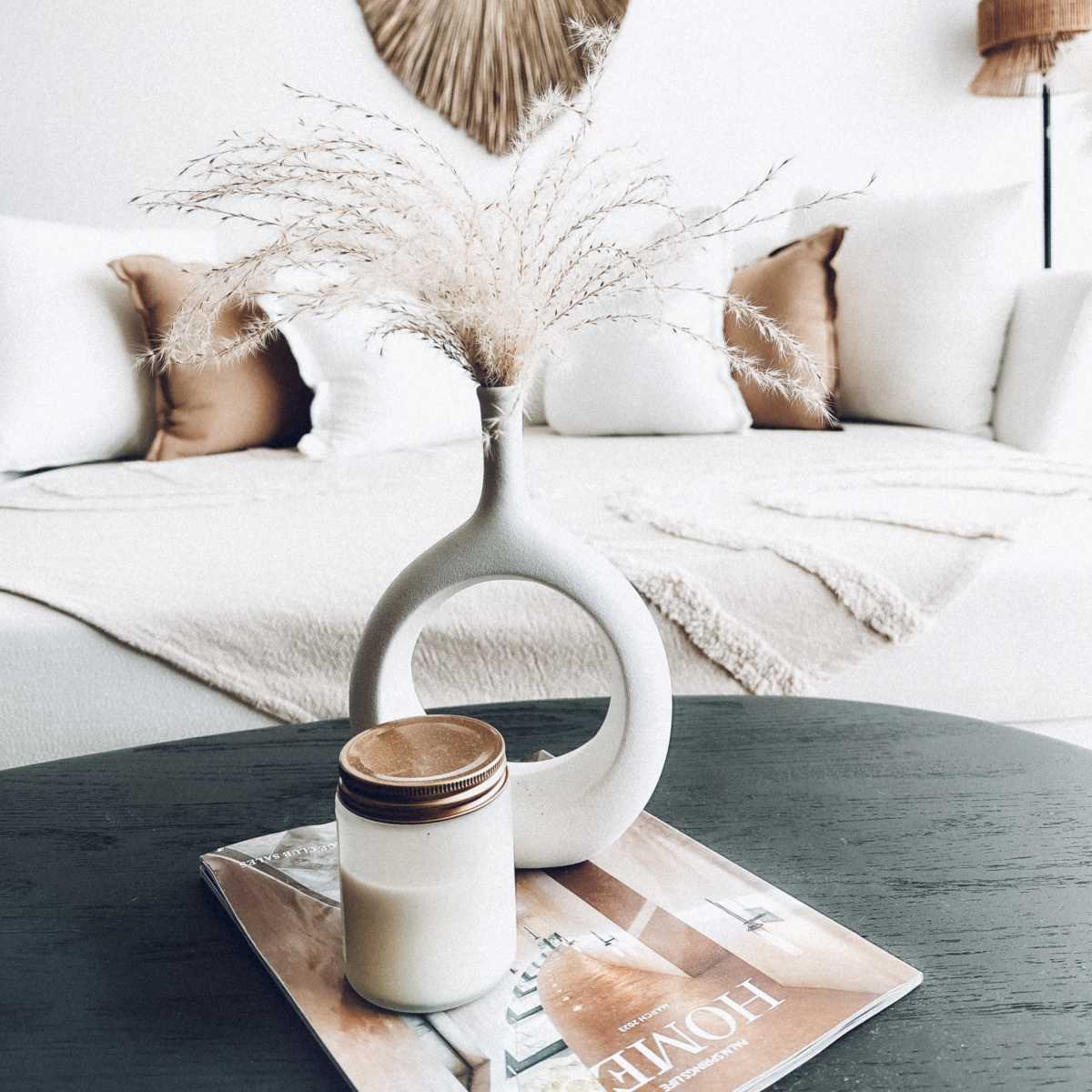 A minimalist living room features a white vase with dried flowers on a dark table, with a magazine and a candle beside it, and decorated cushions and a wall hanging in the background.