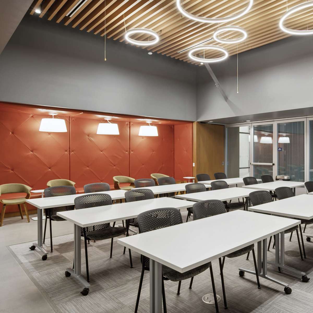 A modern classroom set up with multiple rows of white tables and black chairs, illuminated by circular light fixtures and featuring a red accent wall.