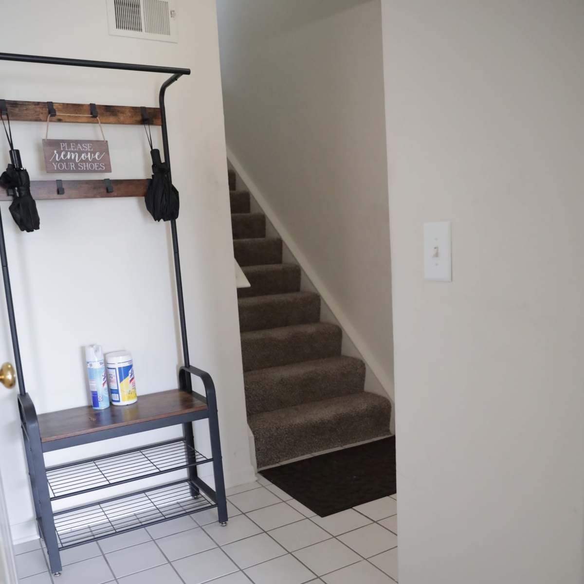 A hallway with a staircase leading upward, a bench rack for coats on the left, and two mats on the tiled floor.