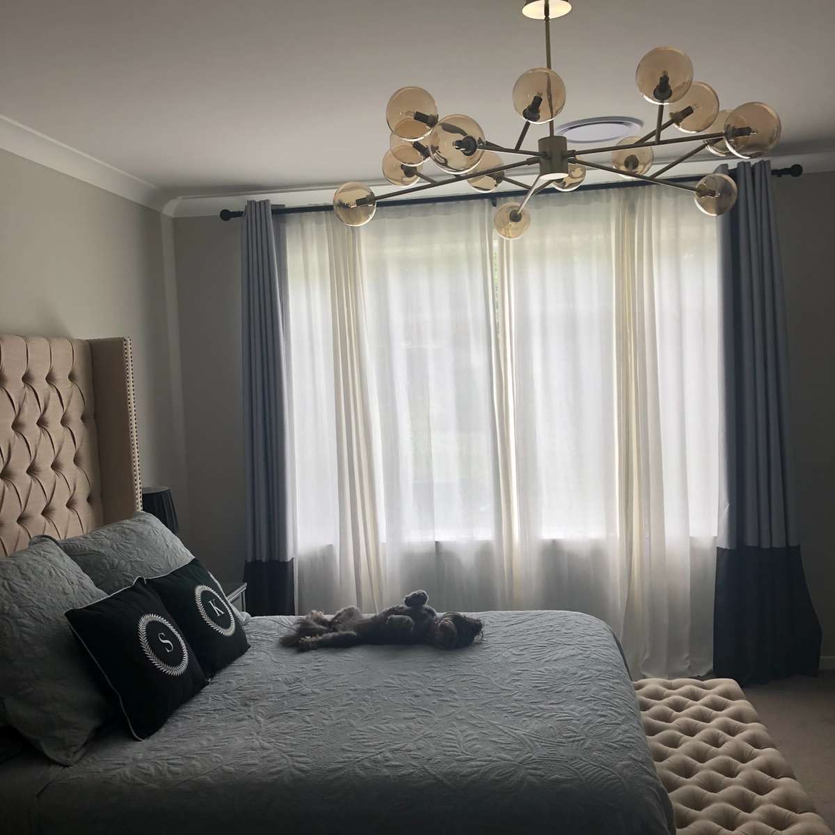 A well-decorated bedroom featuring a large bed with a tufted headboard, a chandelier, and long curtains framing a window.