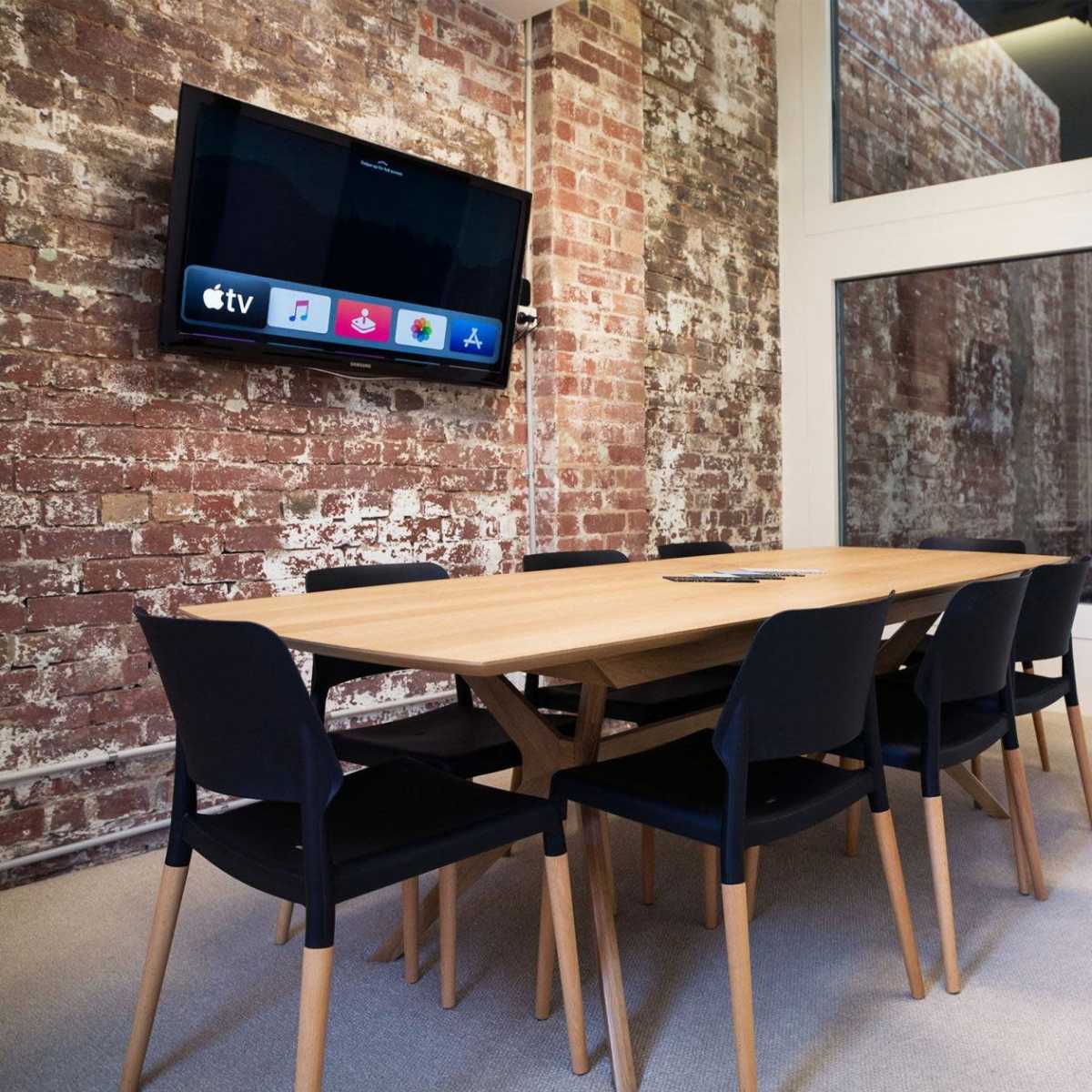 A modern meeting room with a wooden table surrounded by black chairs, a television mounted on a brick wall, and large windows letting in natural light.