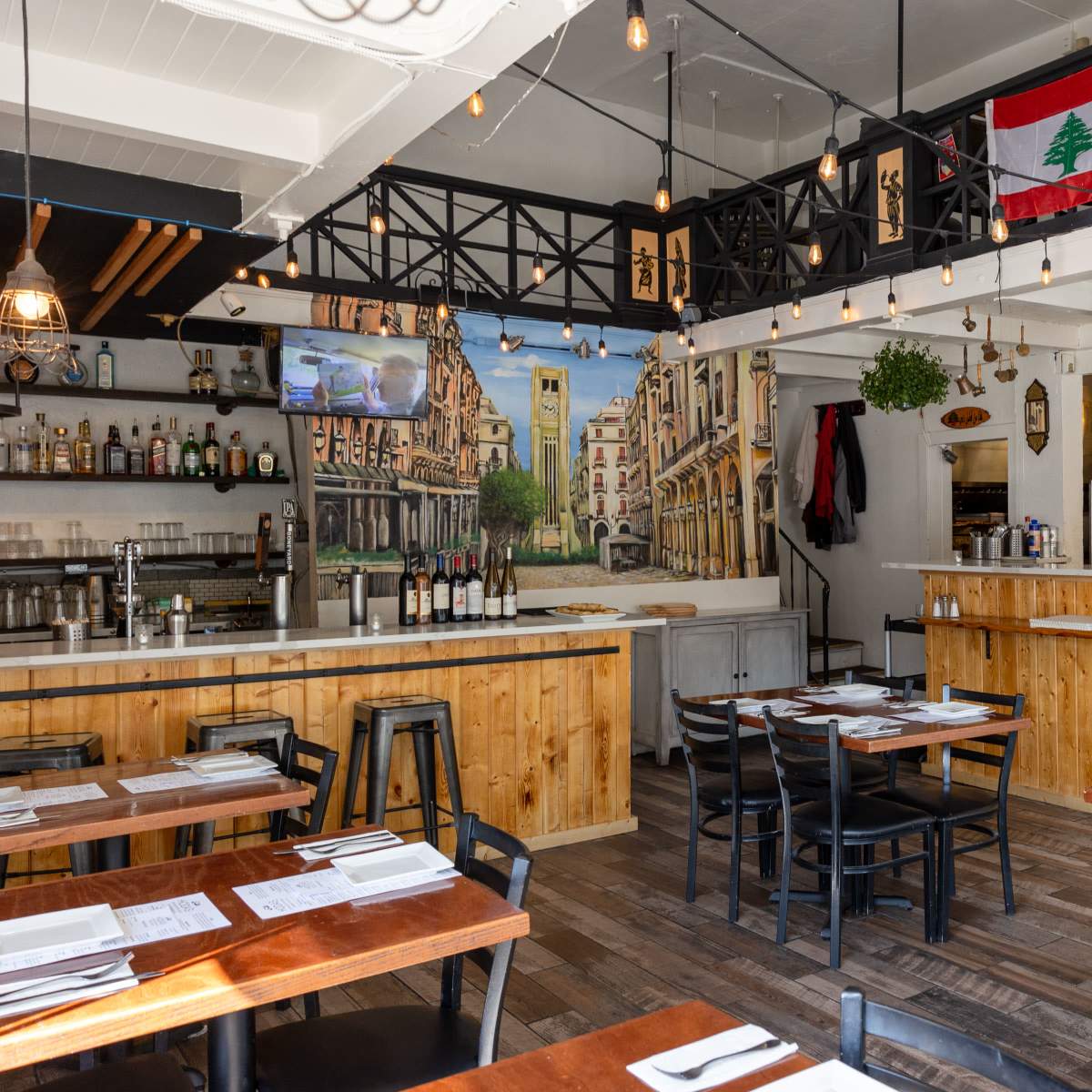 The interior of a restaurant features wooden tables and chairs, a bar area with bottles, and a mural of a street scene, complemented by a Lebanese flag hanging above.