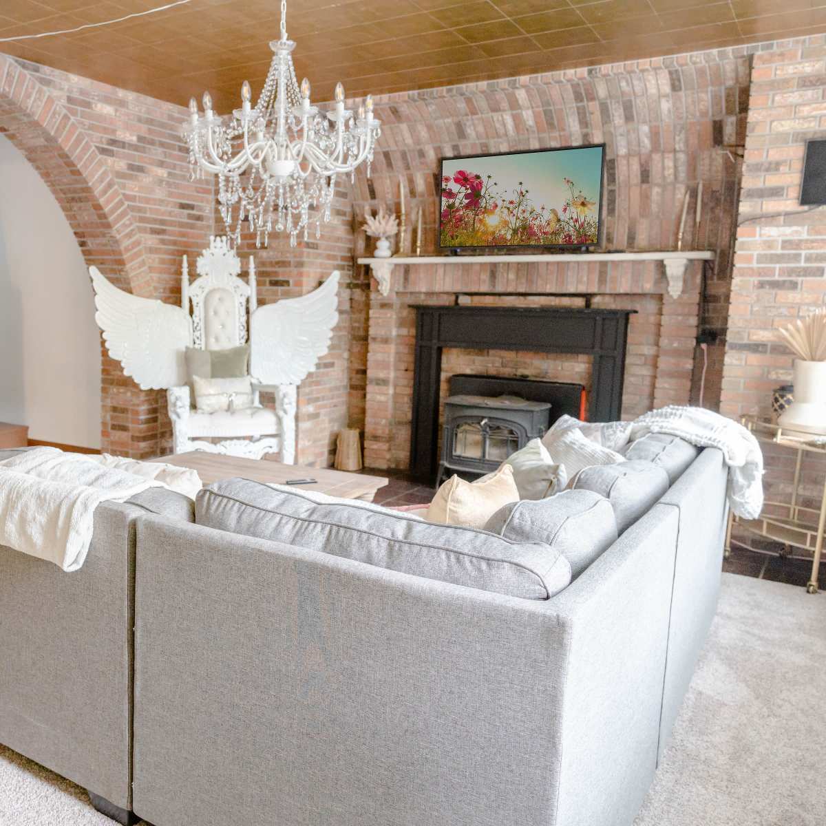 A living room with a gray sectional sofa, a brick fireplace, an ornate chandelier, and a staircase leading to a second level.
