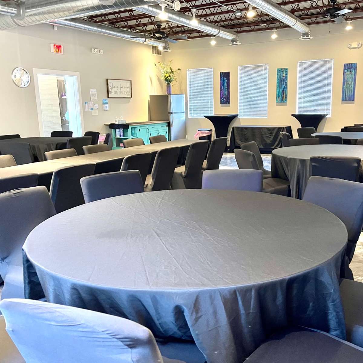 A spacious event room features several round tables covered with black tablecloths, surrounded by dark chairs.