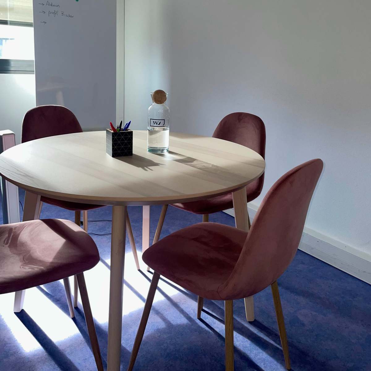 A small meeting room features a round wooden table surrounded by four pink chairs, with a glass water bottle and a pen holder on the table.