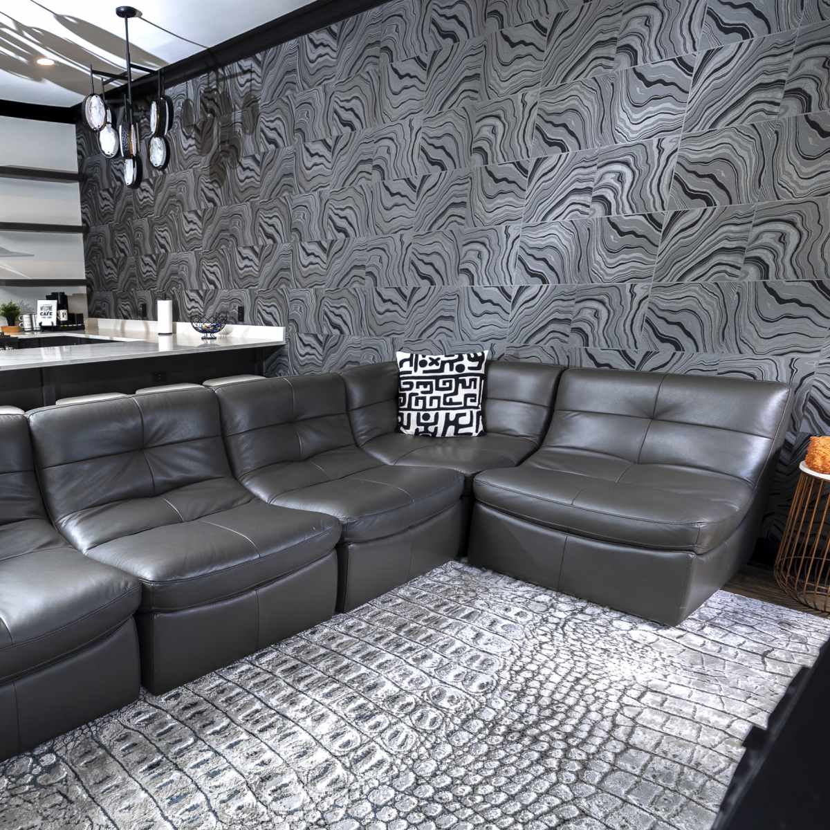 A modern living room with a dark gray leather sectional sofa, patterned throw pillows, and a patterned rug, alongside a sleek kitchen with a stainless steel oven and minimalist cabinetry.
