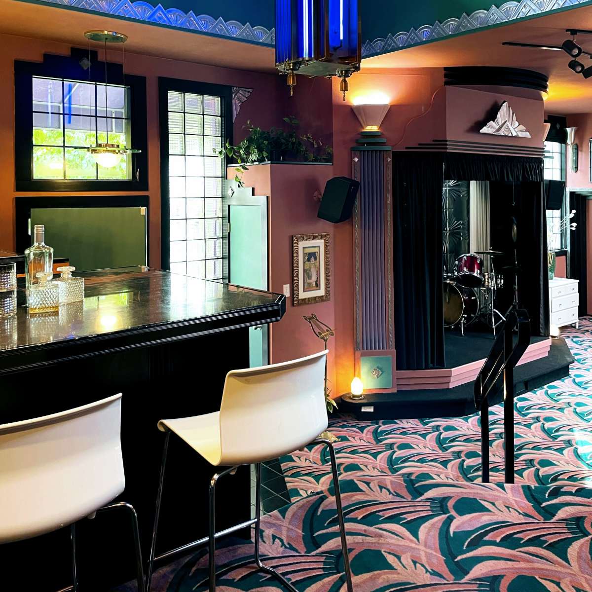 An interior view of a stylish bar featuring a black countertop, white bar stools, and colorful artwork on the walls, with a stage for live performances in the background.