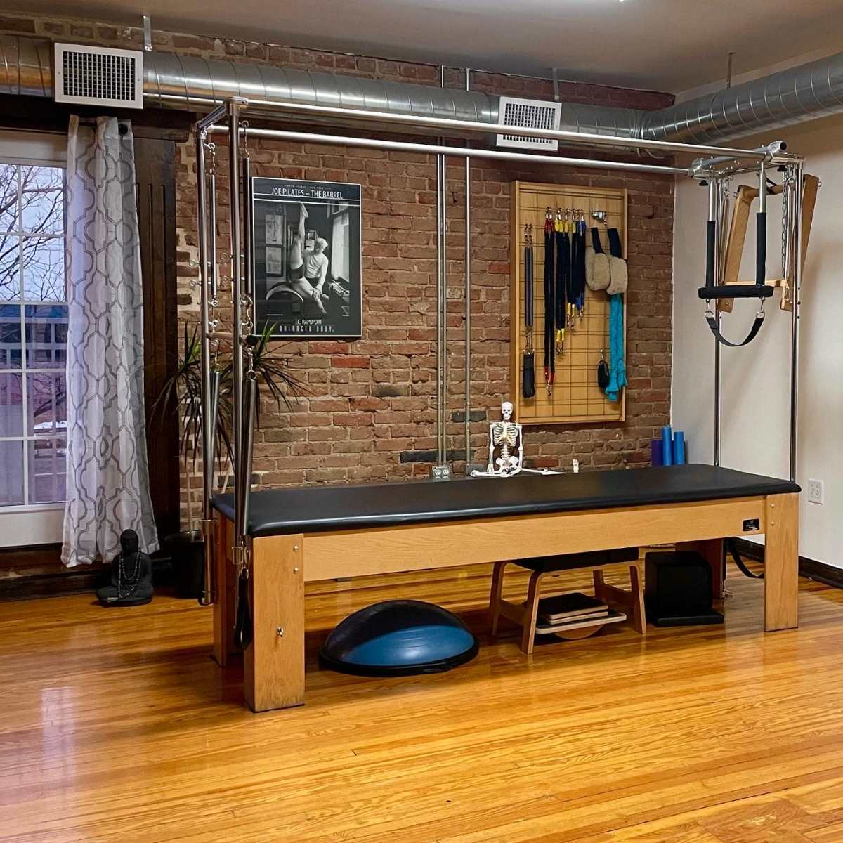 A Pilates reformer machine set up in a studio with wooden floors and brick walls, featuring exercise equipment and a plant nearby.
