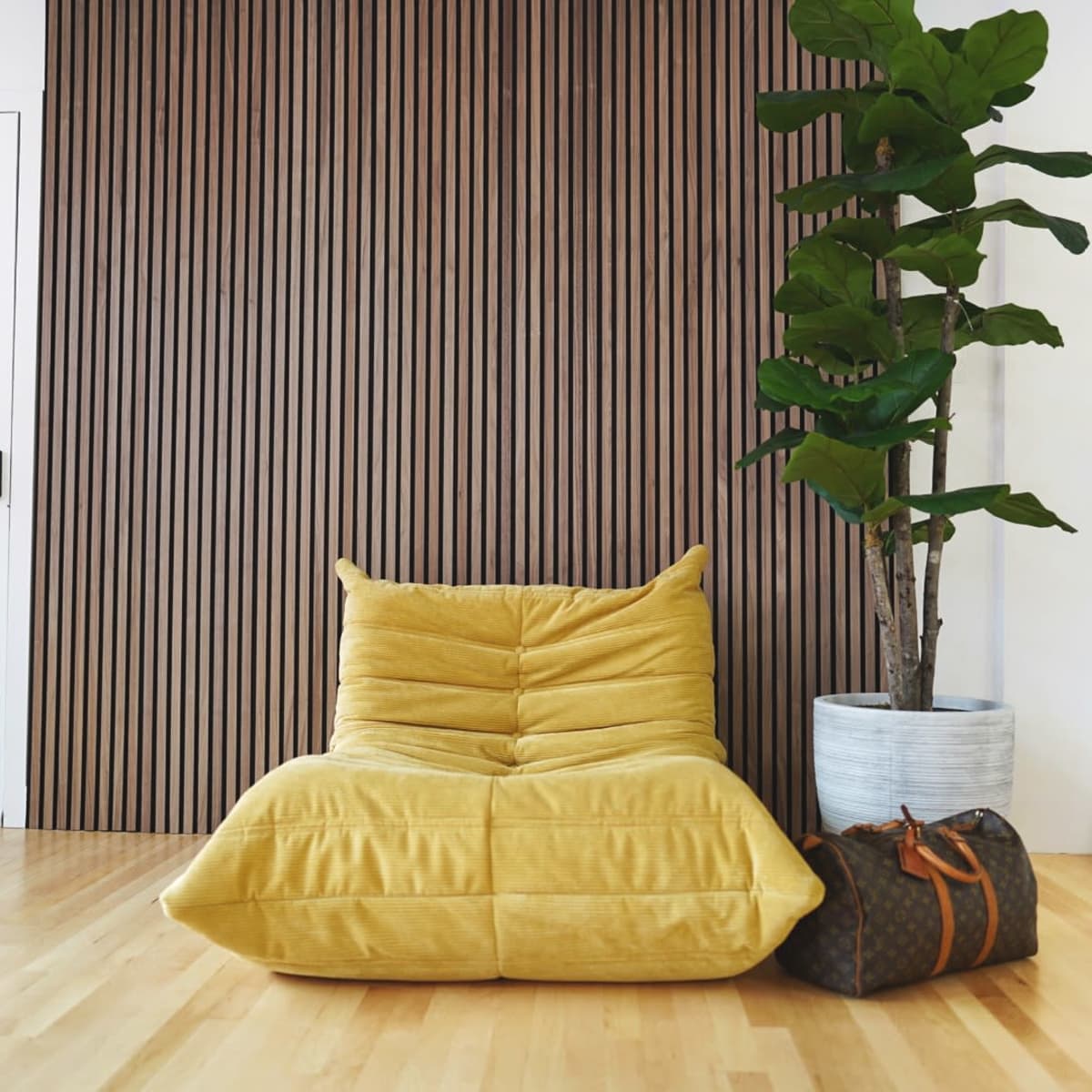 A yellow lounge chair and a potted plant are positioned next to a brown-striped wall and a closed white door in a hardwood-floored room.