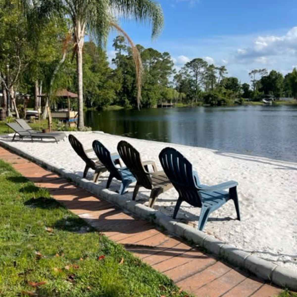 A row of Adirondack chairs faces a calm lake surrounded by trees, with a sandy beach area in the foreground.