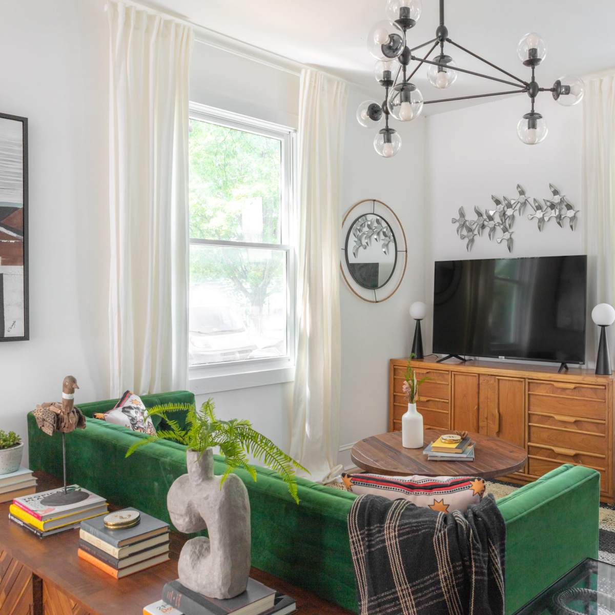 A bright living room featuring a green sofa, a wooden TV cabinet, and large windows with white curtains.