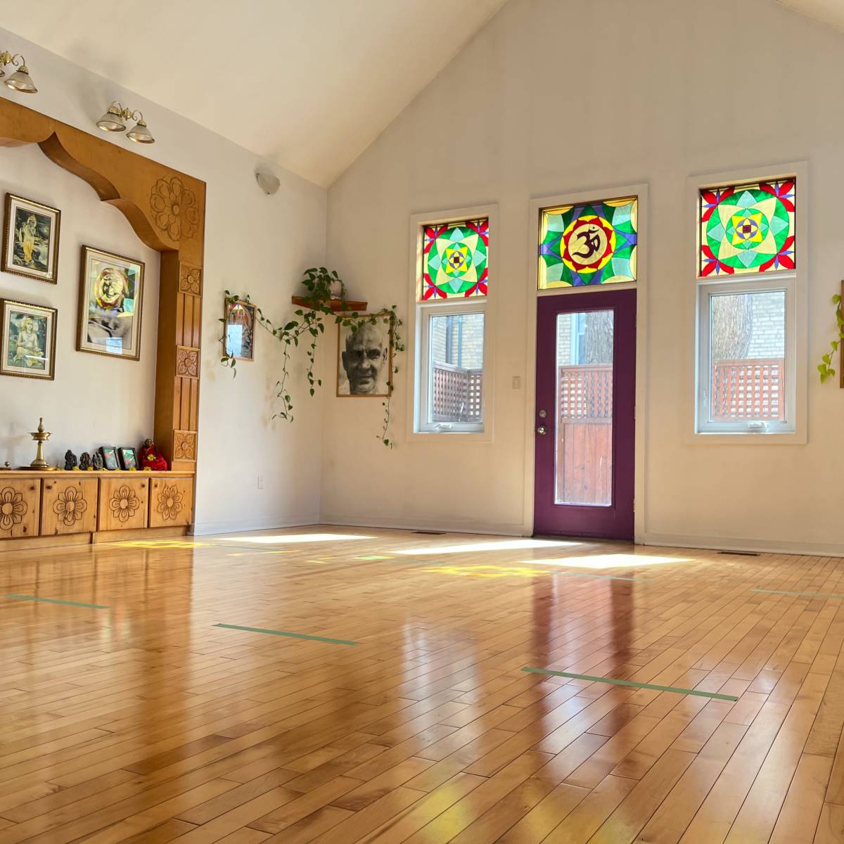 A spacious, well-lit room with wooden flooring, featuring colorful stained glass windows and a decorative wooden wall with framed photographs.