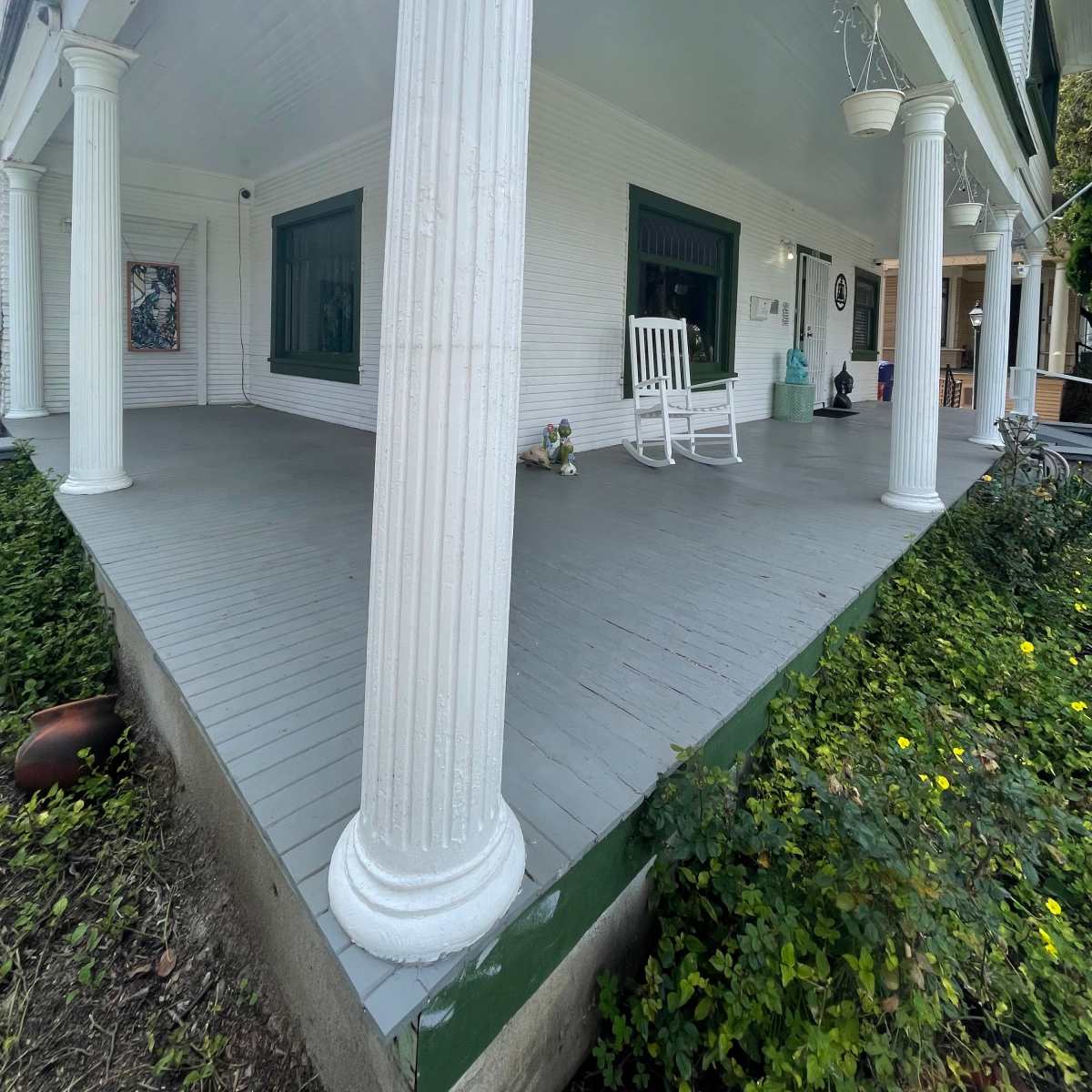 A large porch with white columns, a gray floor, and a rocking chair, surrounded by green foliage.