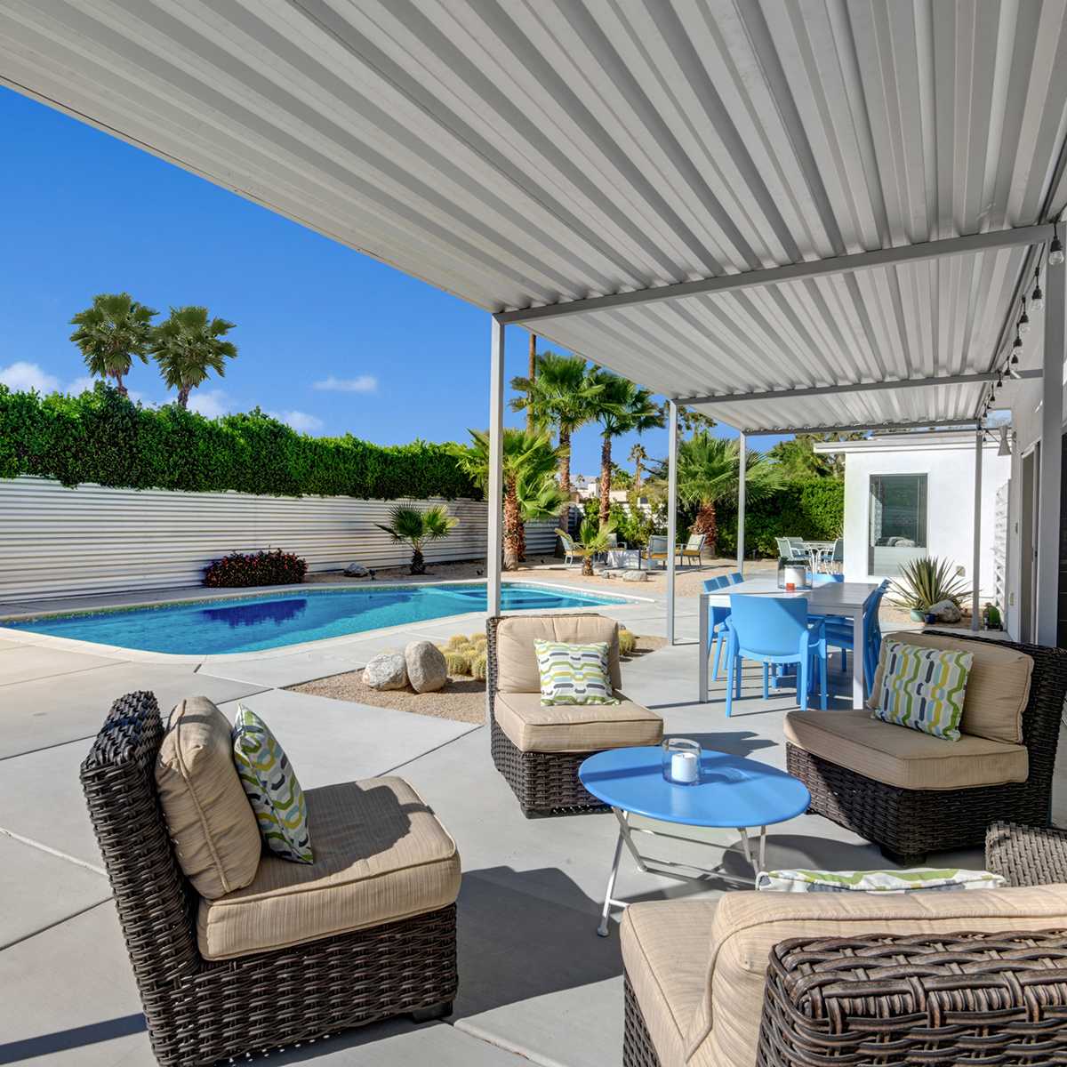 A patio features comfortable seating arrangements and a swimming pool, surrounded by greenery and a bright sky.