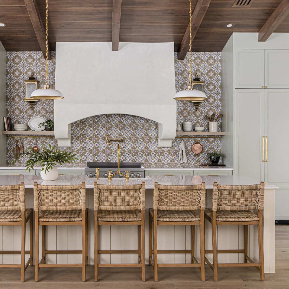 The kitchen features a large central island with woven bar stools, surrounded by light green cabinetry and intricate tile backsplash, complemented by a decorative range hood.
