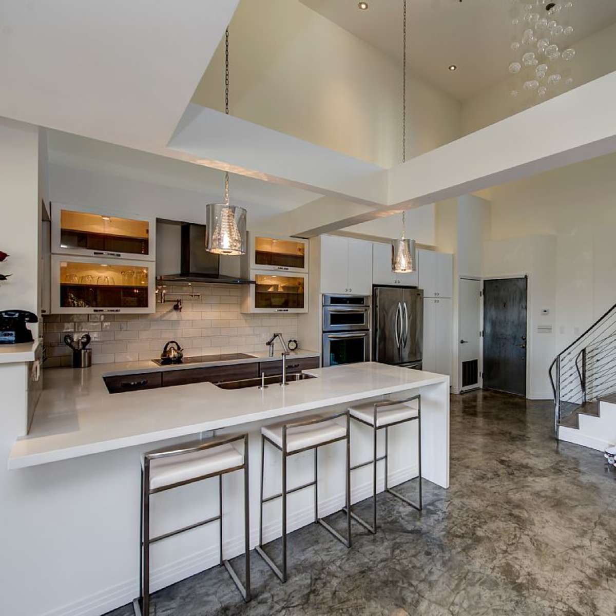 A modern kitchen featuring a large island with two bar stools, stainless steel appliances, and an adjacent staircase with a metallic railing.