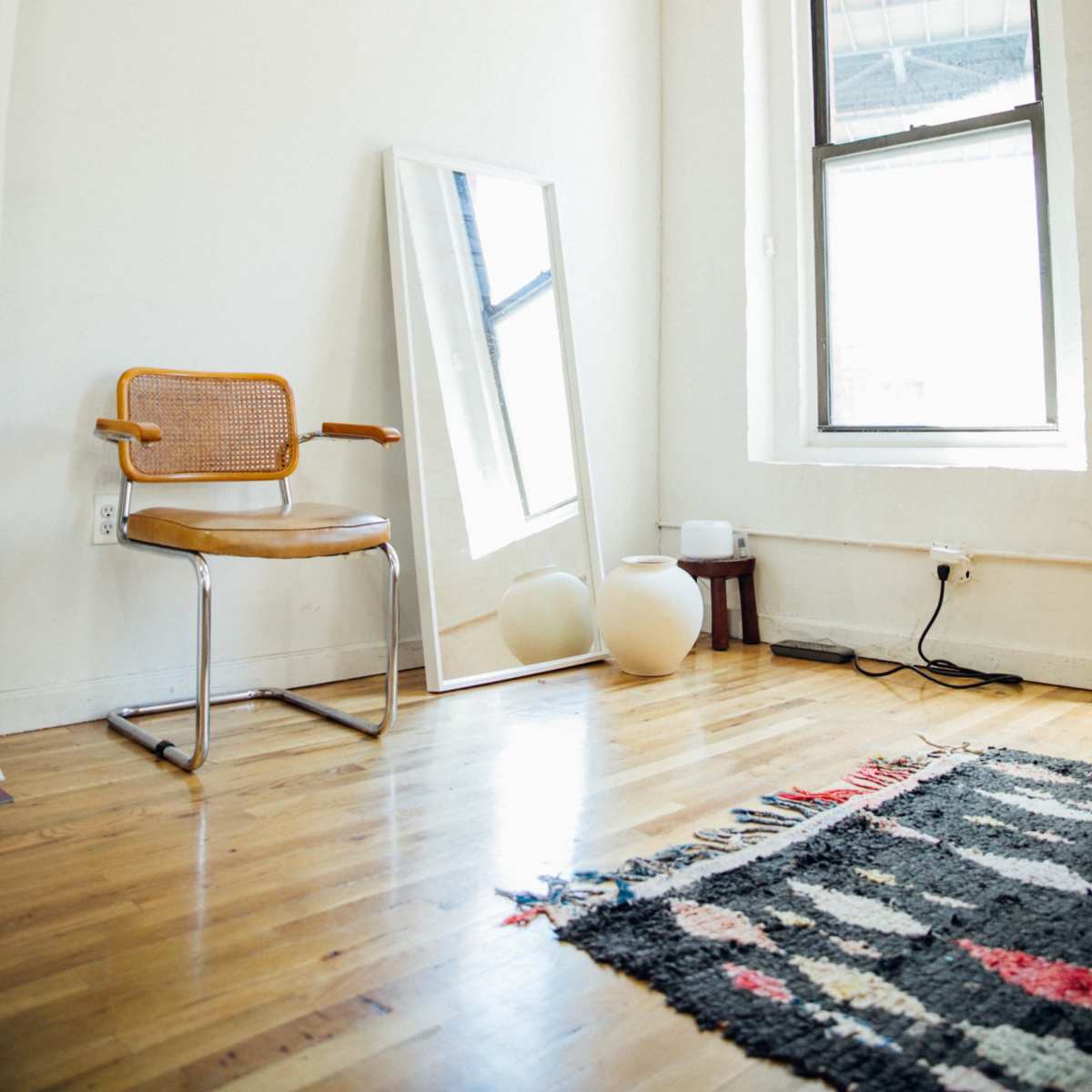 A minimalist room featuring a wooden floor, a chair, a large mirror, two round objects, and a window with exposed brick walls.