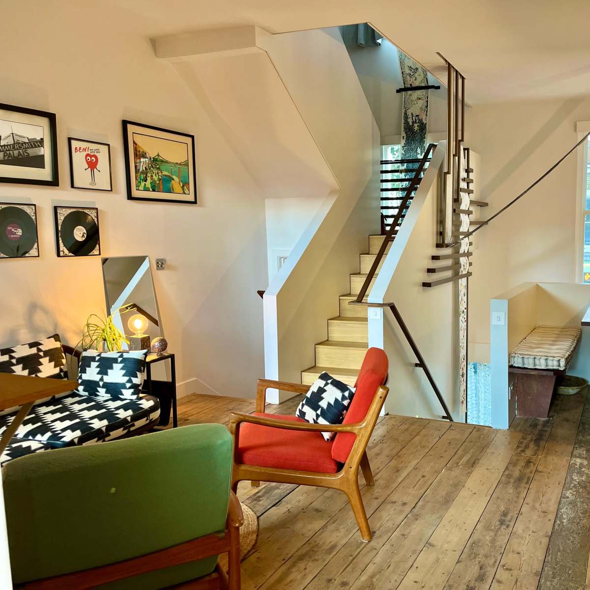 A modern interior space with wooden flooring, a staircase leading to an upper level, a dining table, and a seating area featuring patterned chairs and framed artwork.