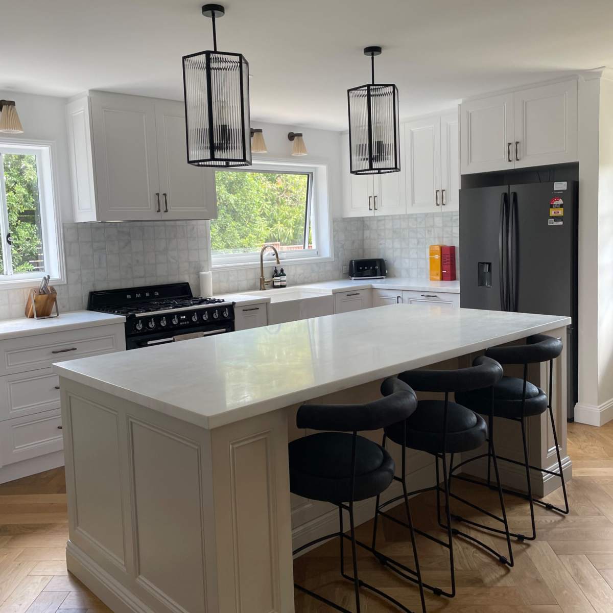 A modern kitchen features a large island with four black stools, white cabinetry, a stainless steel refrigerator, and pendant lighting.