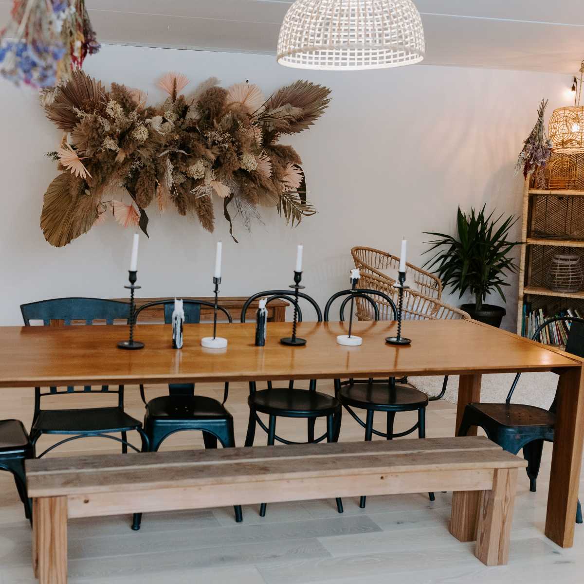 A wooden dining table with black chairs and a bench is set in a well-lit room featuring a decorative wall arrangement of dried plants.