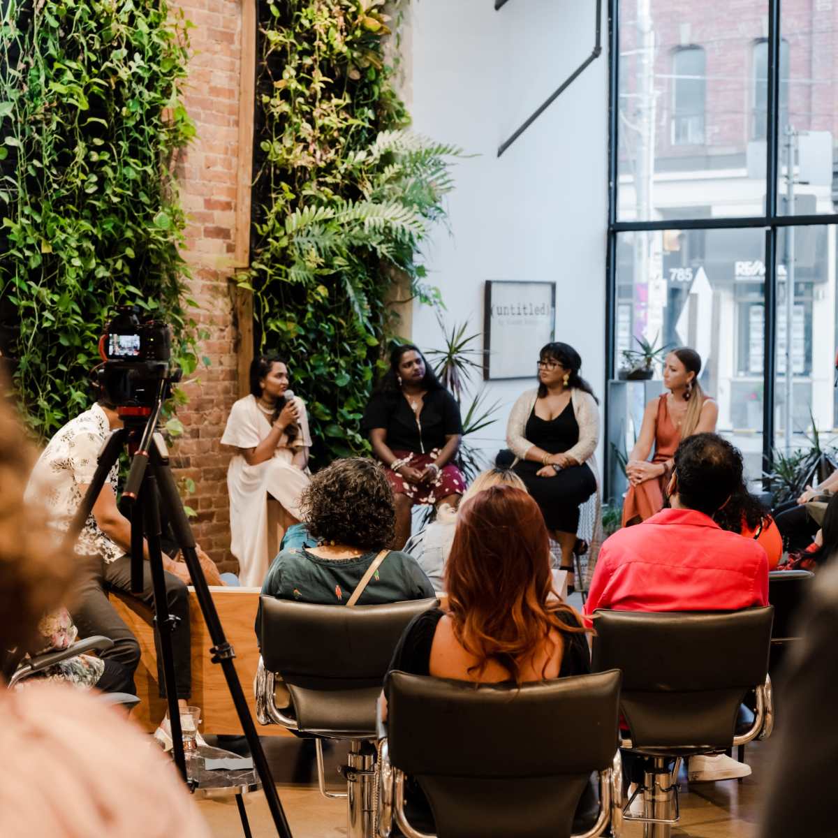 A group of six women participates in a panel discussion in a spacious room with large windows and greenery on the walls, while an audience listens in chairs.