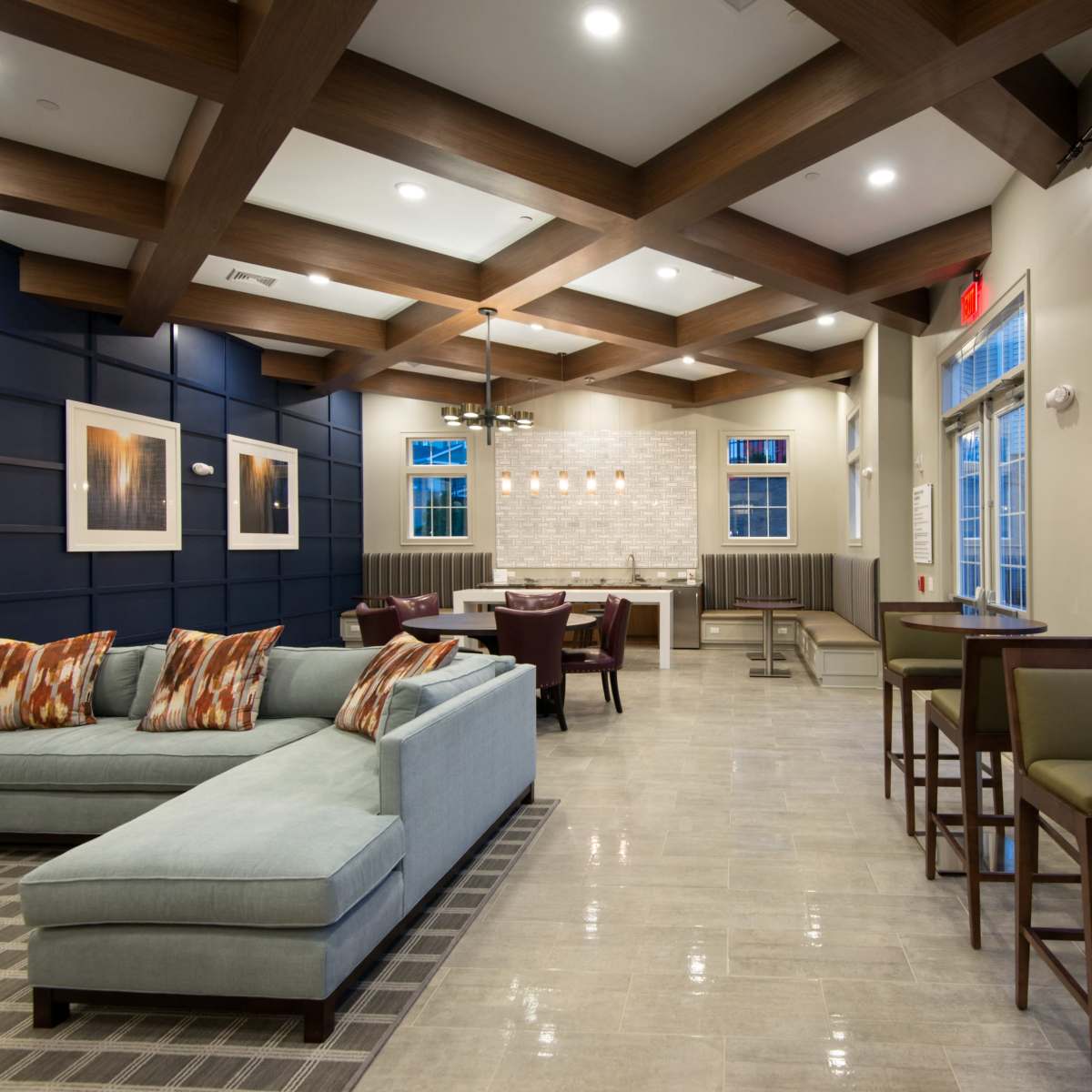 A modern common area with a blue sectional sofa, wooden beams on the ceiling, and a small dining space with high chairs and tables.