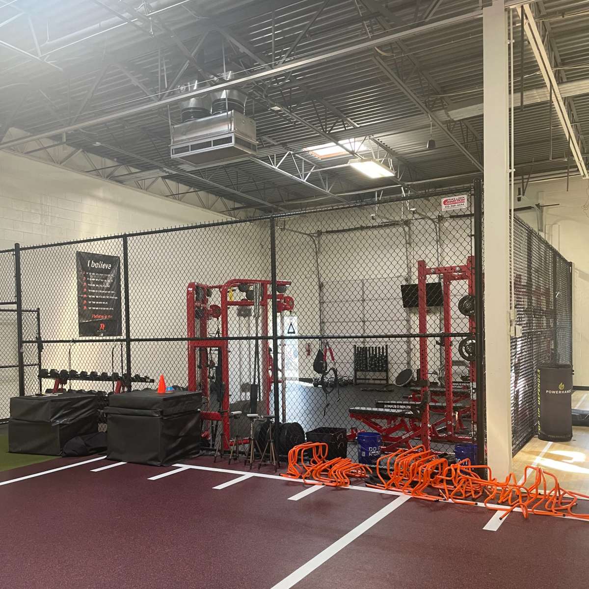 A gym area enclosed with a chain-link fence, featuring weightlifting equipment and training tools along with a rubberized floor.
