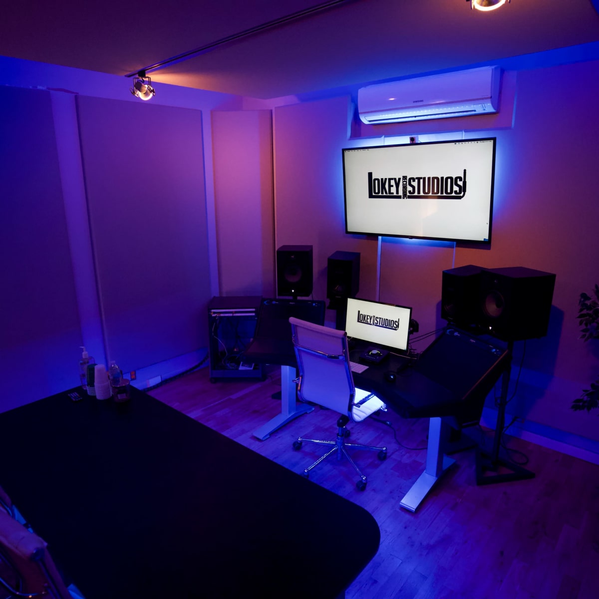 A modern recording studio with a desk, computer, and large speakers, illuminated by blue lighting.