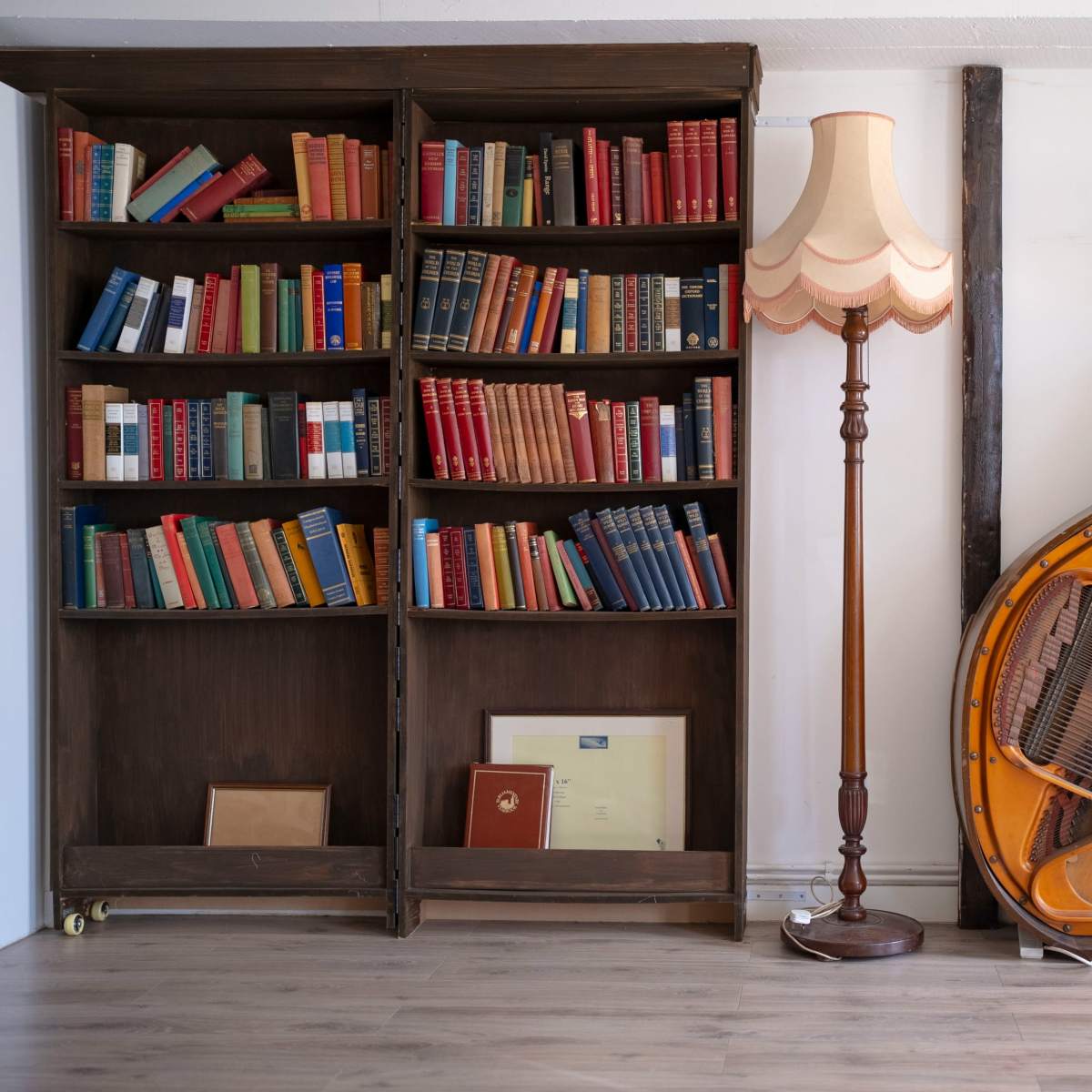 A wooden bookshelf filled with colorful books stands next to a vintage lamp and a grand piano.