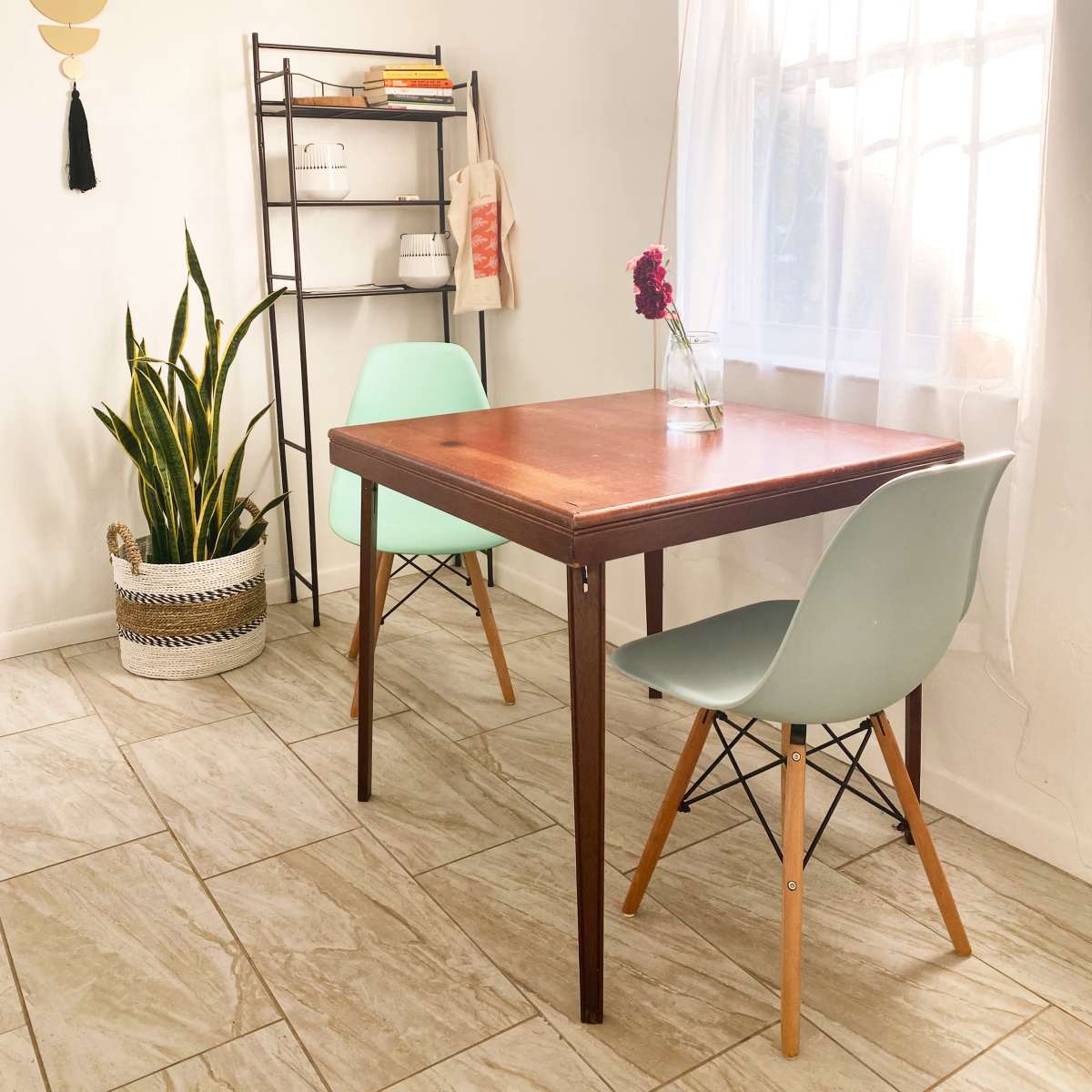 A wooden table is set between two light green chairs in a room featuring a large window, a plant in a woven pot, and a bookshelf in the background.