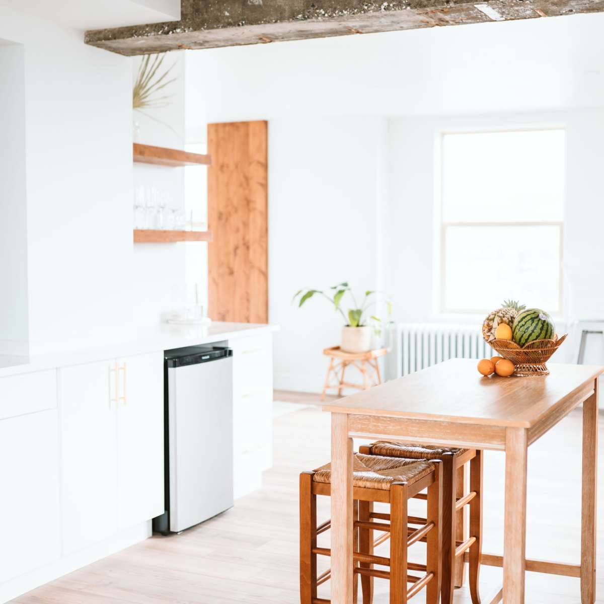 A bright kitchen features a wooden table surrounded by stools, with a fruit bowl atop the table and modern cabinetry in the background.