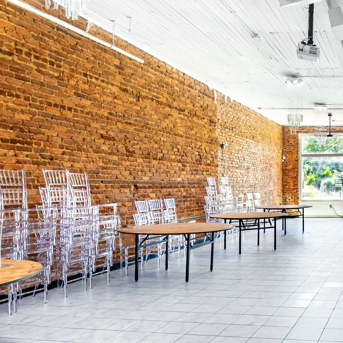 A spacious event room with exposed brick walls, stacked clear chairs, and wooden tables arranged in the area.