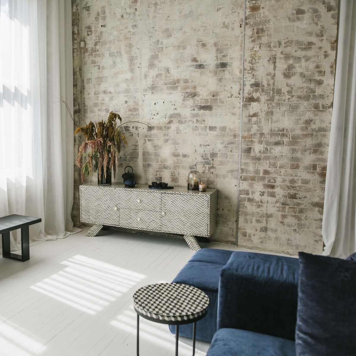 A minimalist living room features a textured brick wall, a dark blue sofa, a geometric-patterned sideboard, and a small circular table.
