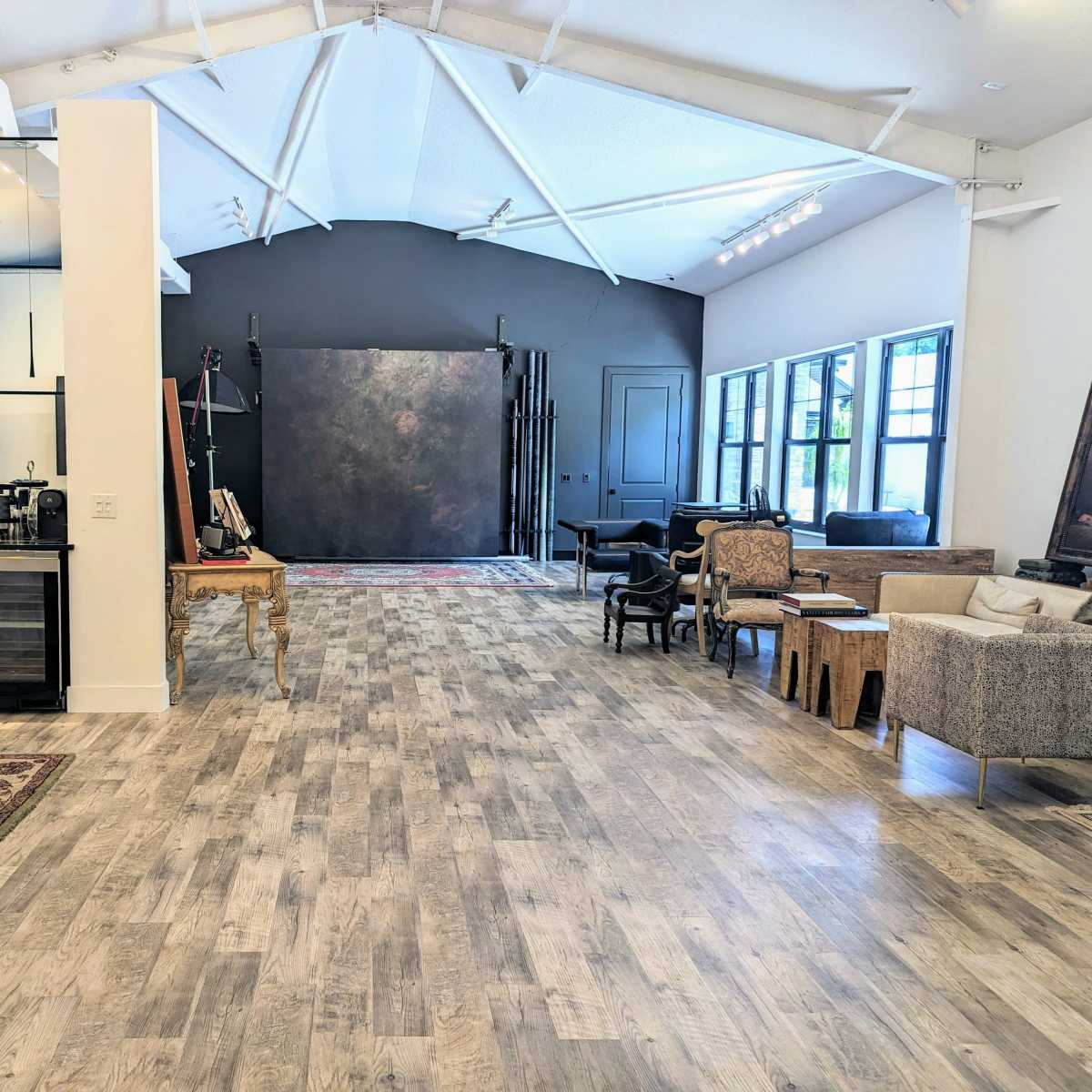 A spacious room with a modern kitchen area on one side and a stylish seating arrangement on the other, featuring a mix of furniture styles and large windows allowing natural light in.