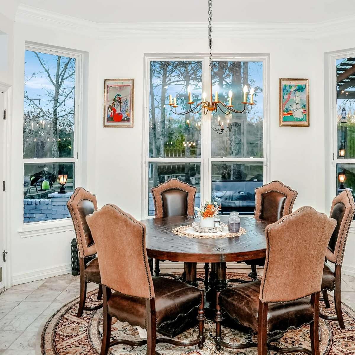 A round wooden dining table surrounded by six upholstered chairs is situated in a bright, airy dining area with large windows and decorative artwork on the walls.