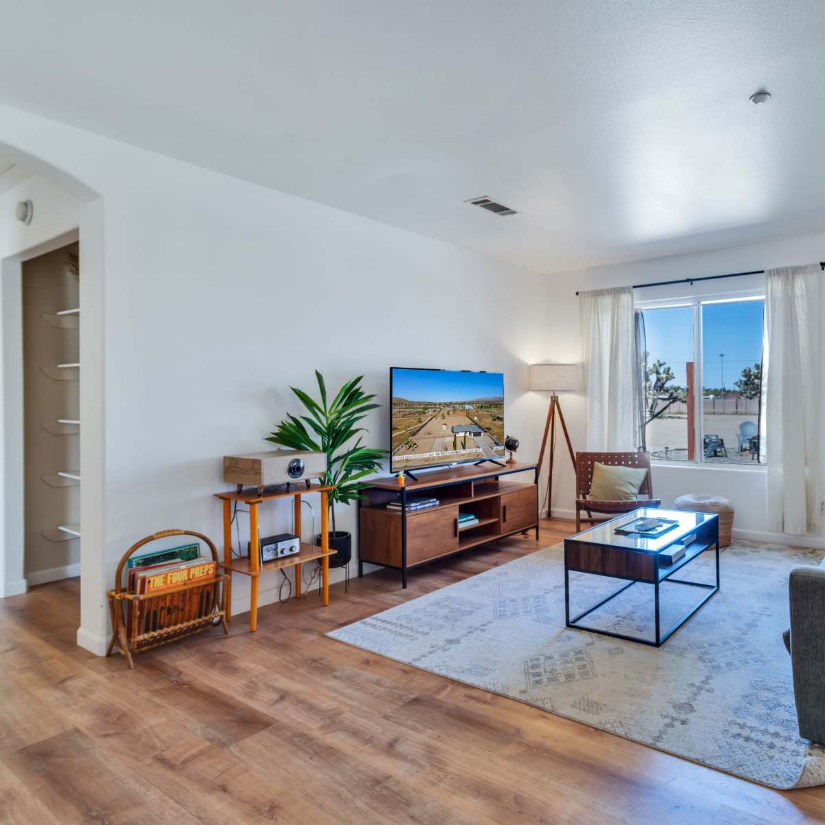 A modern living room features a large television, a comfortable sofa, a coffee table, and a window with sheer curtains allowing natural light.