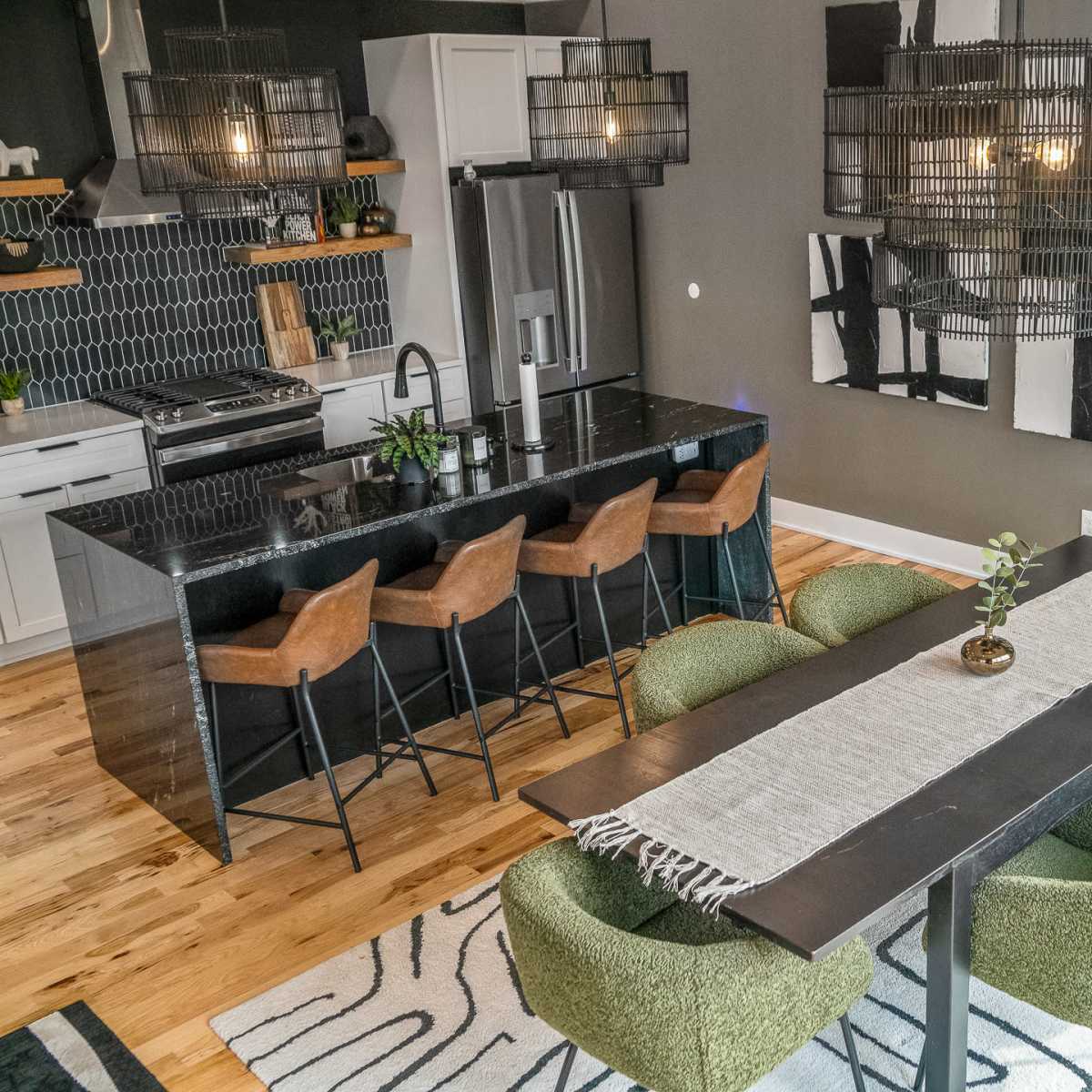 A modern dining area features a long black table surrounded by green chairs, with a bar and kitchen visible in the background, complemented by pendant lights and abstract wall art.