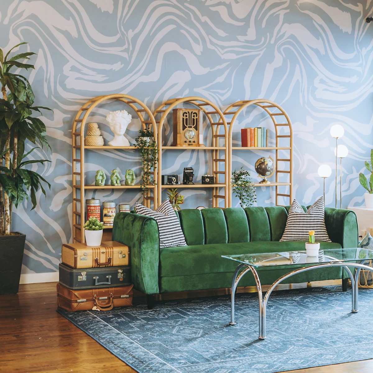 A green velvet sofa is placed in front of a large window with a blue abstract wall design, accompanied by a glass coffee table and a bookshelf filled with decorative items and plants.
