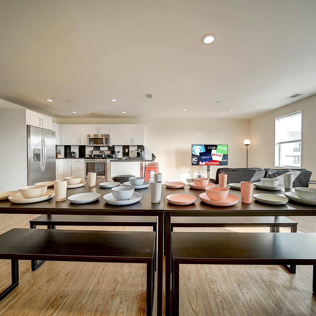 A dining table is set with various plates and cups, positioned in a modern kitchen and living area with light-colored walls and wooden flooring.