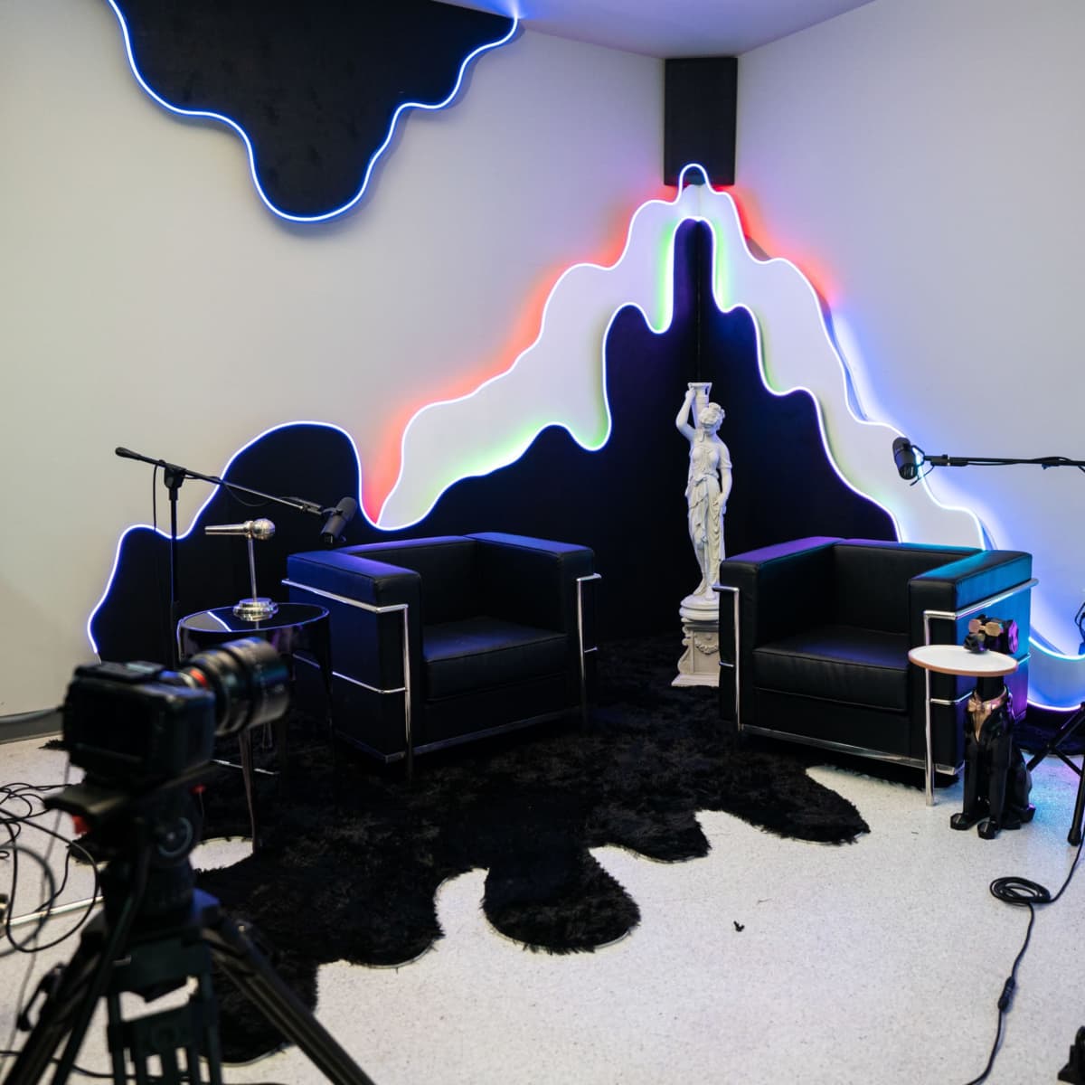 A modern interview set features two black chairs, a statue, and colorful neon lighting against a stark wall, with cameras positioned for filming.