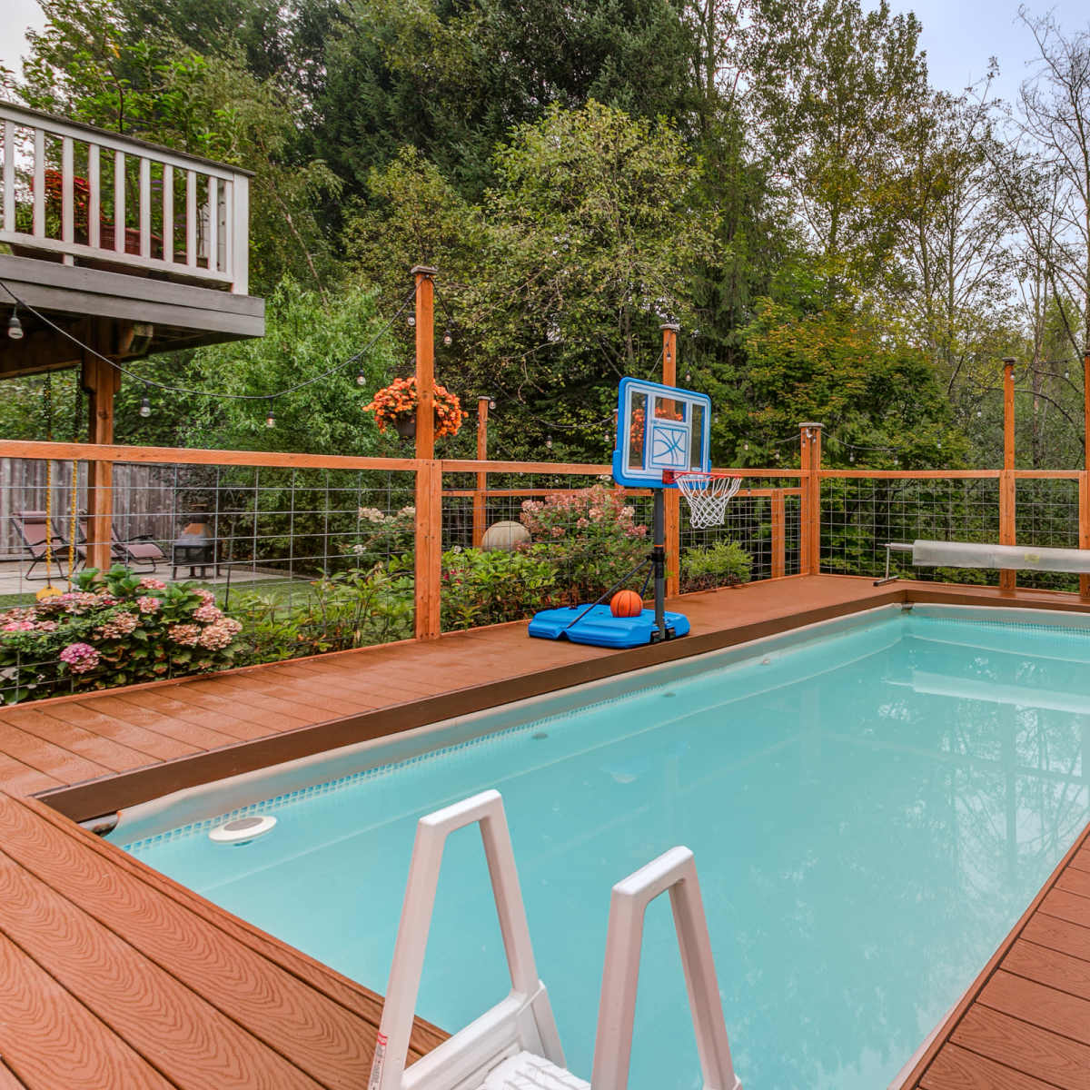 A backyard with a swimming pool surrounded by a wooden deck, a basketball hoop, and lush greenery in the background.