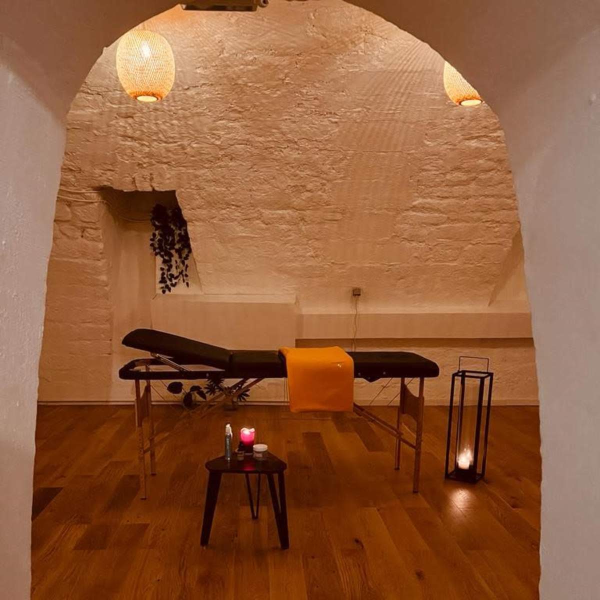 A massage room features a treatment table with an orange towel, surrounded by candles and wooden flooring.