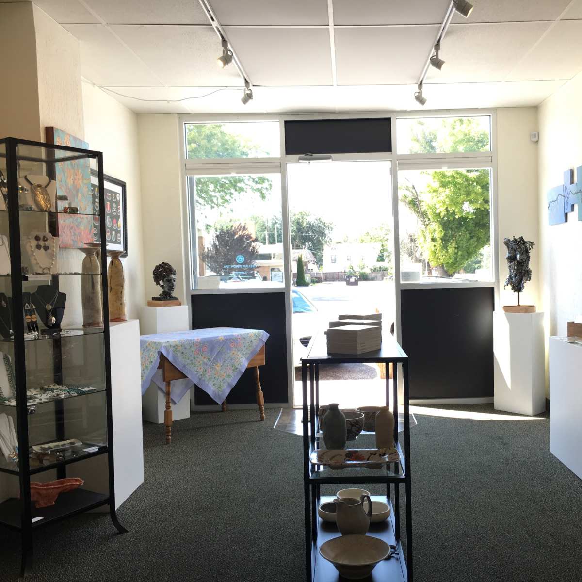 An art gallery interior with display cases and tables showcasing various artworks and crafts, illuminated by natural light from large windows.