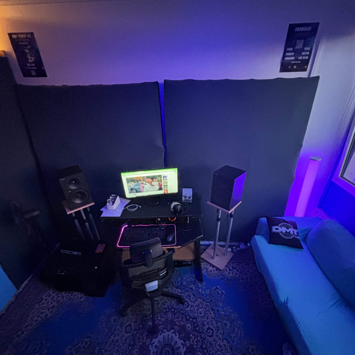 A small room features a desk with a computer, speakers, and a chair, surrounded by soundproofing panels and illuminated by soft purple lighting.
